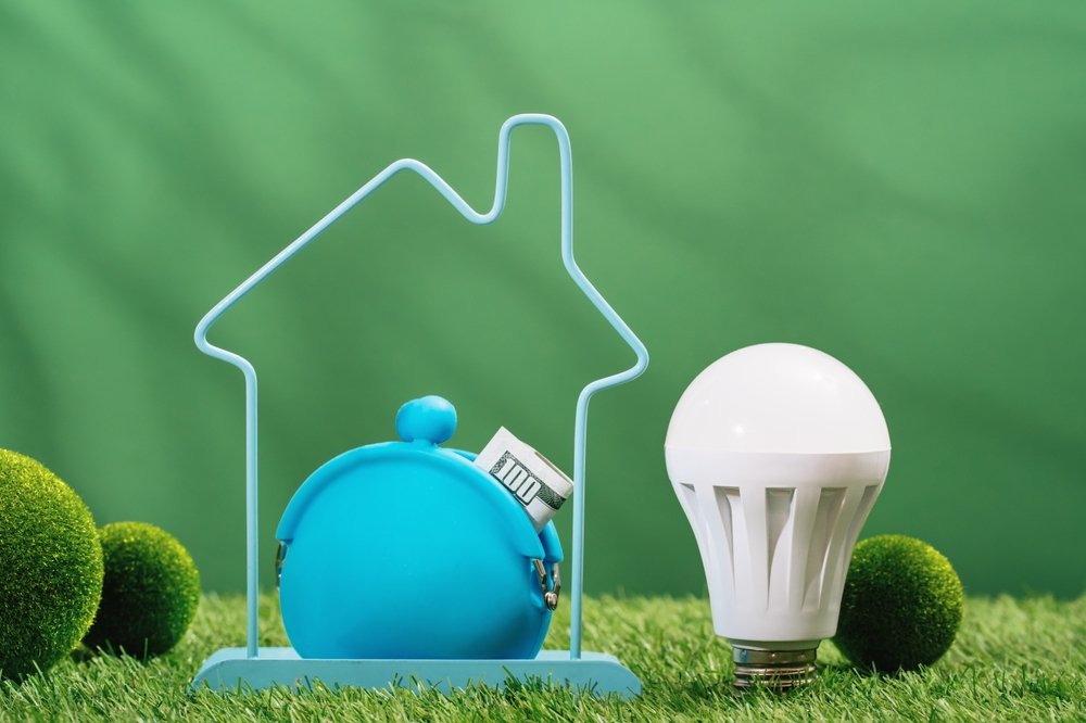 Energy-efficient LED bulb next to a piggy bank within a house outline, symbolizing cost savings with energy-efficient lighting.