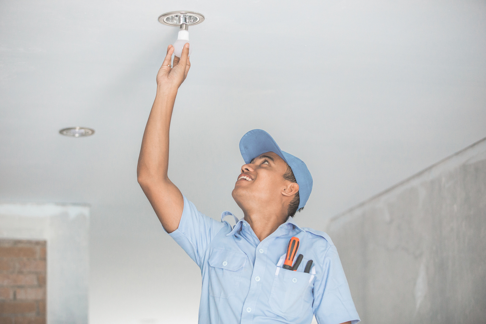 Electrician in blue uniform reaching to install an LED light bulb in a ceiling socket.